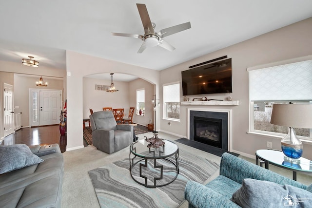 living area featuring baseboards, a fireplace with flush hearth, carpet floors, ceiling fan with notable chandelier, and arched walkways