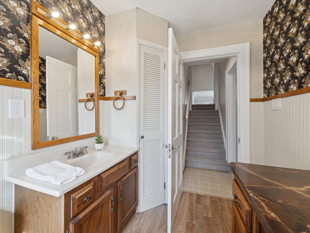 bathroom with vanity, wood finished floors, wainscoting, and wallpapered walls