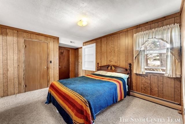 carpeted bedroom with a baseboard heating unit, wooden walls, visible vents, and a textured ceiling