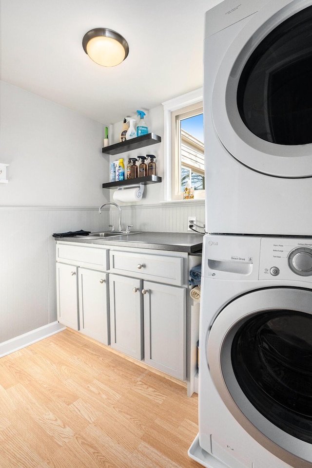 clothes washing area with a wainscoted wall, cabinet space, a sink, light wood-style floors, and stacked washer and clothes dryer