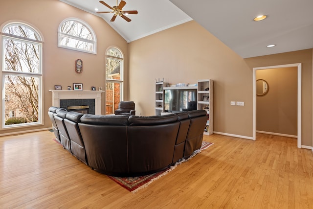 living room with light wood-type flooring, high vaulted ceiling, a ceiling fan, a tiled fireplace, and baseboards