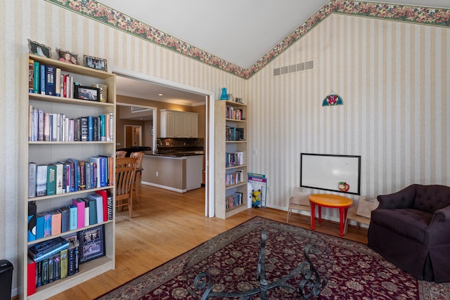 living area with visible vents, lofted ceiling, wood finished floors, and wallpapered walls