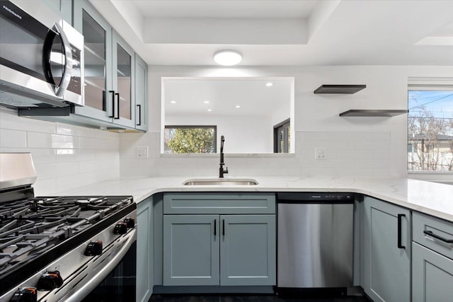 kitchen with a sink, glass insert cabinets, appliances with stainless steel finishes, decorative backsplash, and light stone countertops