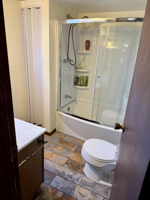 bathroom featuring baseboards, bath / shower combo with glass door, toilet, stone tile floors, and vanity