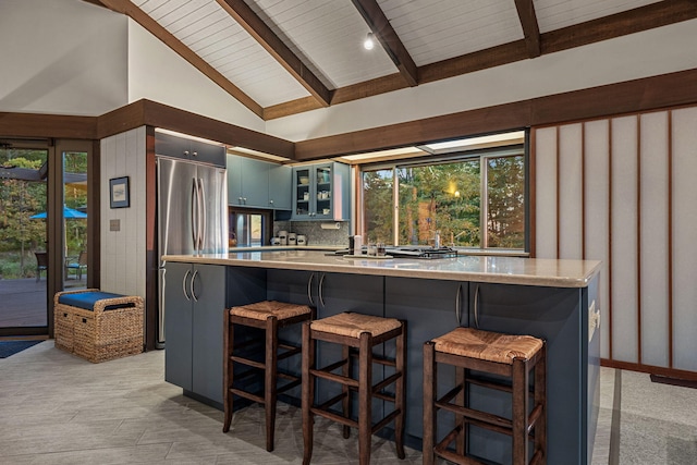 kitchen featuring stainless steel refrigerator, lofted ceiling with beams, a kitchen breakfast bar, tasteful backsplash, and glass insert cabinets