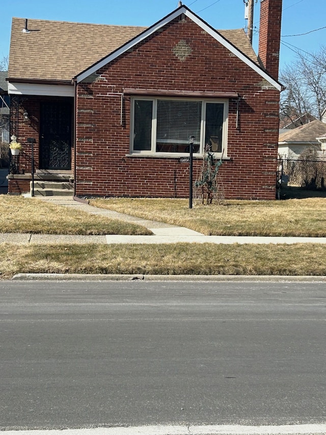 view of front of house featuring brick siding, a shingled roof, fence, a front yard, and a chimney