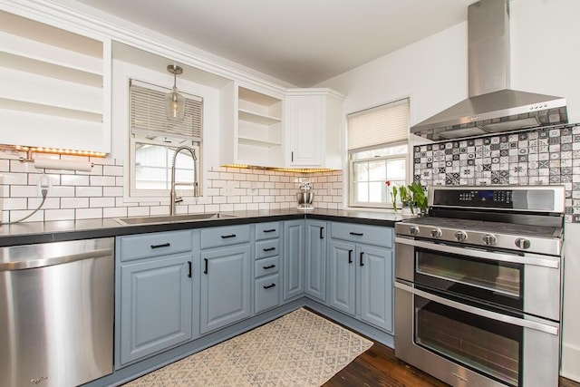 kitchen with dark countertops, open shelves, stainless steel appliances, wall chimney exhaust hood, and a sink