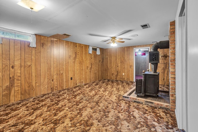 basement featuring visible vents, ceiling fan, wood walls, carpet floors, and a wood stove