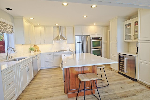 kitchen with beverage cooler, an island with sink, a sink, stainless steel appliances, and wall chimney range hood