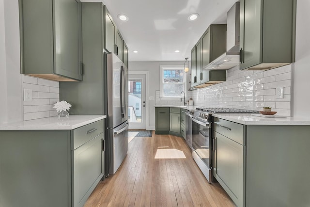 kitchen featuring light wood-type flooring, green cabinetry, appliances with stainless steel finishes, wall chimney exhaust hood, and tasteful backsplash