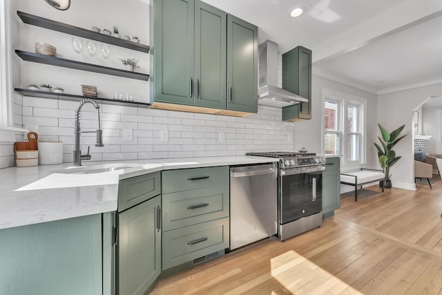 kitchen with green cabinetry, open shelves, a sink, appliances with stainless steel finishes, and wall chimney range hood