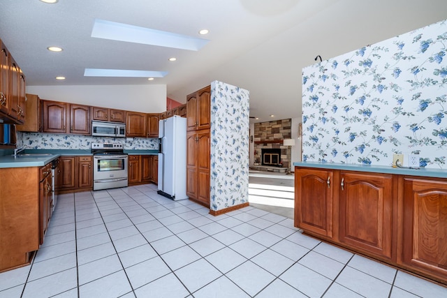 kitchen featuring brown cabinetry, vaulted ceiling with skylight, light tile patterned flooring, a fireplace, and appliances with stainless steel finishes