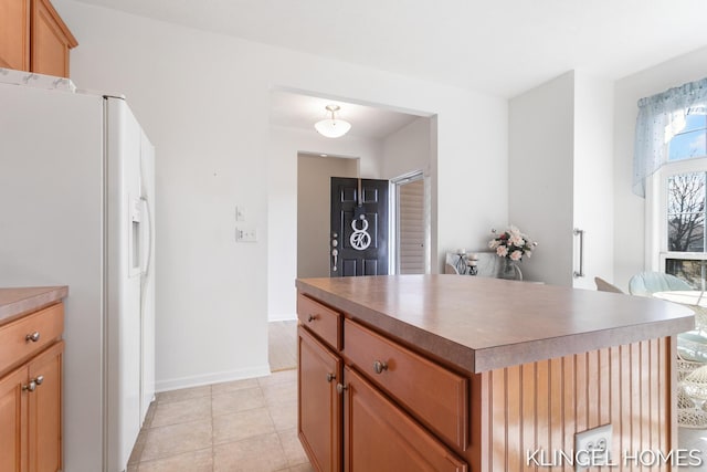 kitchen featuring a kitchen island, white refrigerator with ice dispenser, light tile patterned flooring, brown cabinetry, and baseboards