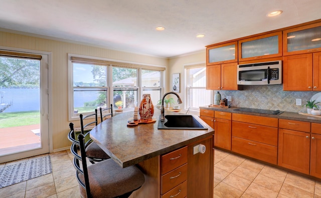 kitchen with an island with sink, a sink, stainless steel microwave, black electric cooktop, and brown cabinets