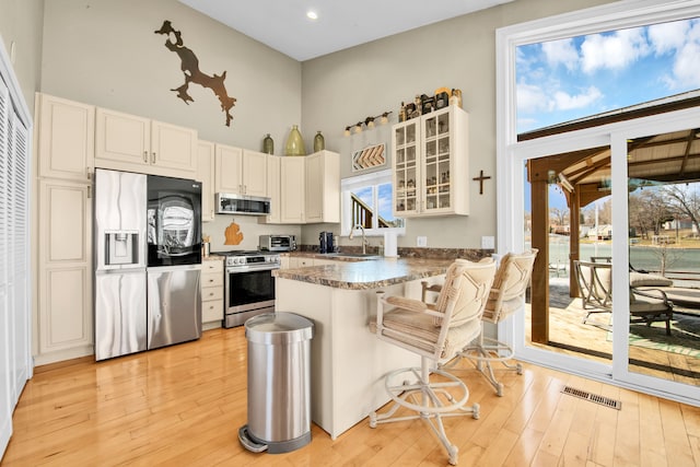 kitchen with a peninsula, light wood-style flooring, a high ceiling, stainless steel appliances, and glass insert cabinets