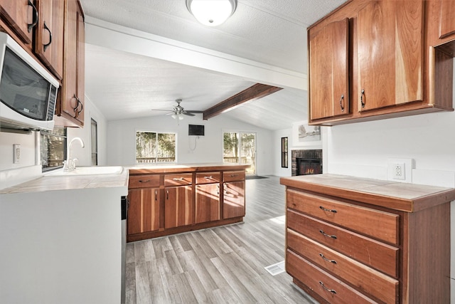 kitchen with stainless steel microwave, vaulted ceiling with beams, tile counters, brown cabinets, and a sink