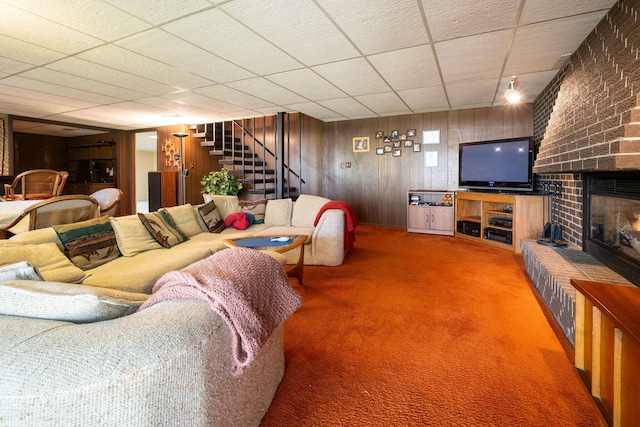 carpeted living area featuring stairs, wooden walls, a fireplace, and a paneled ceiling