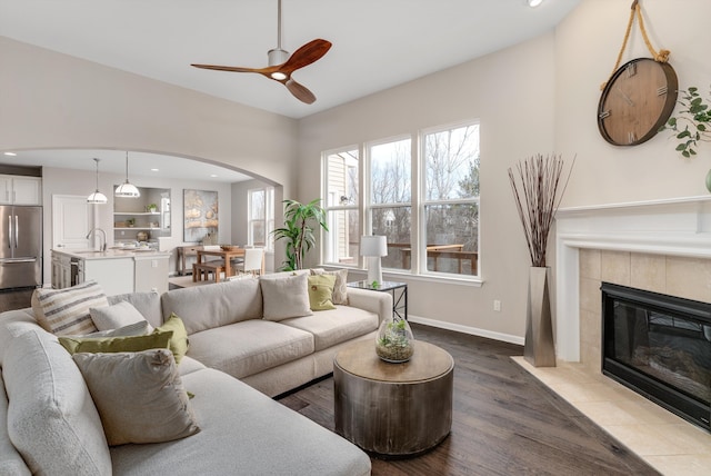 living room with baseboards, a tile fireplace, wood finished floors, arched walkways, and a ceiling fan