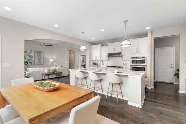 kitchen with tasteful backsplash, under cabinet range hood, double oven, arched walkways, and gas stovetop