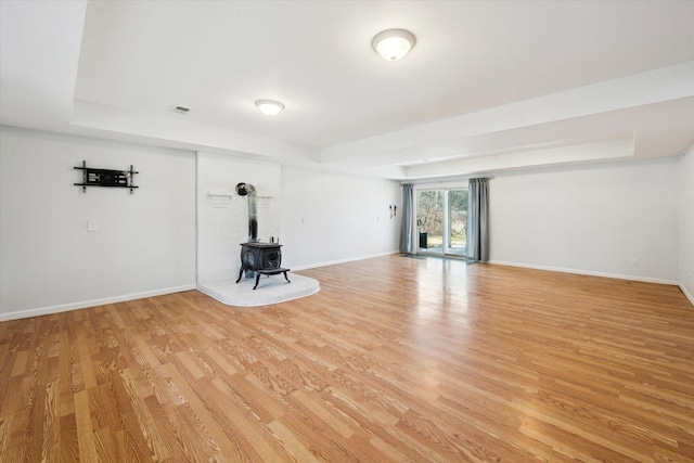 unfurnished living room with baseboards, a raised ceiling, a wood stove, and light wood finished floors