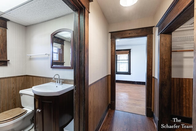 half bath with toilet, wood finished floors, wooden walls, wainscoting, and vanity