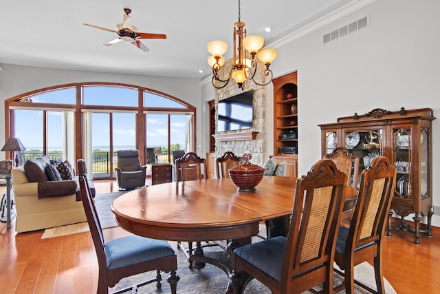 dining room featuring built in features, visible vents, light wood finished floors, a stone fireplace, and ceiling fan with notable chandelier
