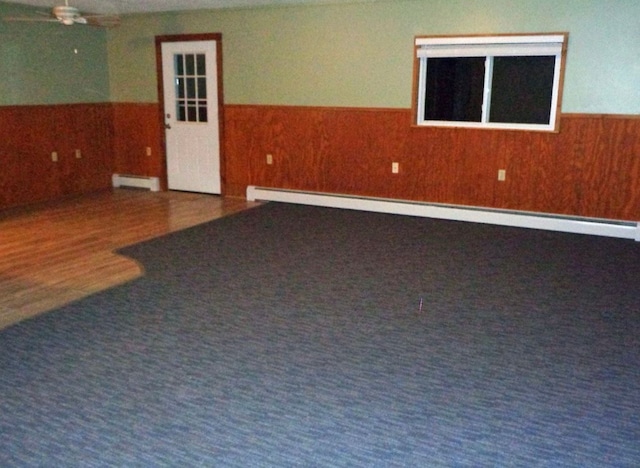 carpeted empty room with a ceiling fan, baseboard heating, wood walls, and wainscoting