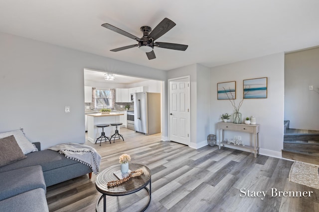 living room featuring light wood-type flooring, baseboards, a ceiling fan, and stairs