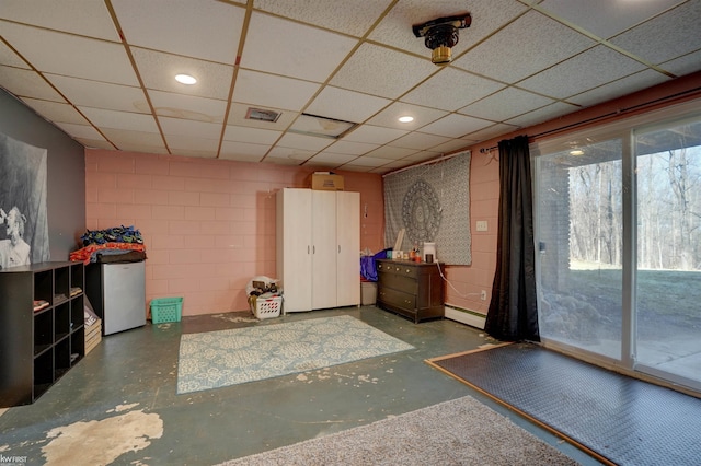 interior space with visible vents, a drop ceiling, a baseboard heating unit, fridge, and concrete block wall