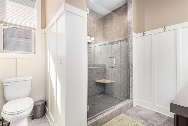 bathroom with a stall shower, stone finish floor, wainscoting, a decorative wall, and toilet