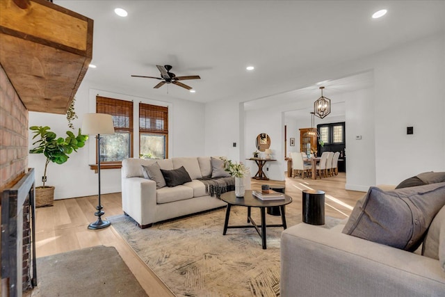 living room with ceiling fan with notable chandelier, a brick fireplace, recessed lighting, and light wood-style floors