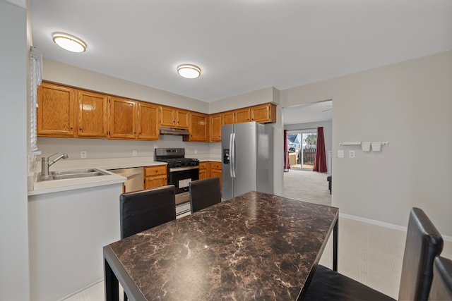 kitchen featuring under cabinet range hood, a sink, stainless steel appliances, brown cabinetry, and baseboards