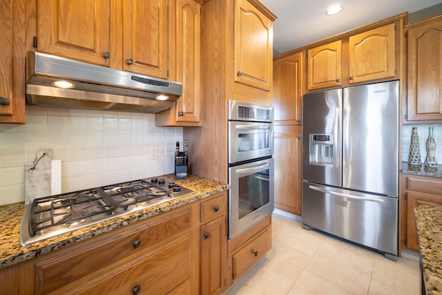 kitchen with under cabinet range hood, light stone countertops, appliances with stainless steel finishes, and brown cabinets