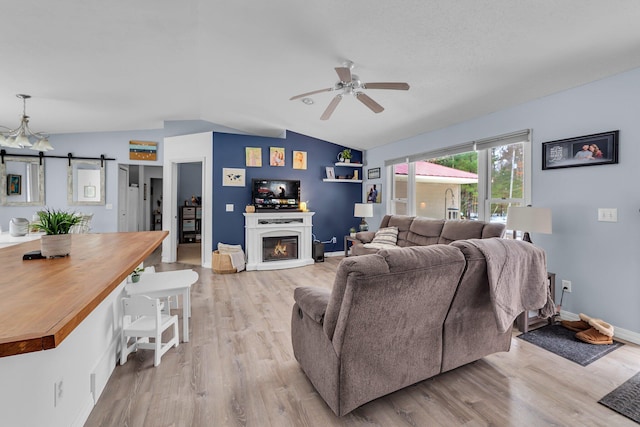 living room featuring light wood-type flooring, a ceiling fan, a glass covered fireplace, a barn door, and vaulted ceiling