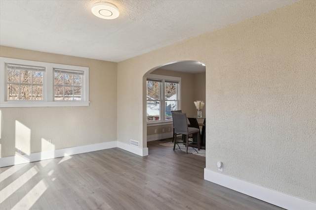 empty room featuring arched walkways, a textured wall, baseboards, and wood finished floors