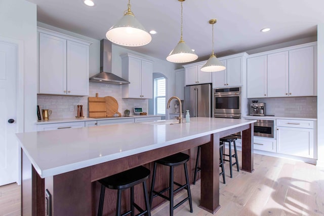 kitchen featuring wall chimney range hood, appliances with stainless steel finishes, light wood-style floors, a large island, and a sink