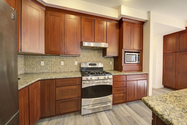 kitchen featuring light stone countertops, stainless steel appliances, light wood-style floors, under cabinet range hood, and tasteful backsplash