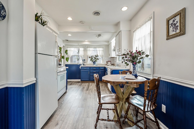 kitchen with blue cabinetry, a wainscoted wall, light wood-type flooring, white appliances, and a sink