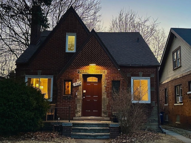 tudor house featuring cooling unit, brick siding, and roof with shingles