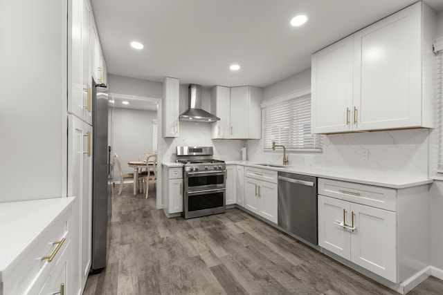 kitchen with wall chimney range hood, wood finished floors, white cabinets, stainless steel appliances, and a sink