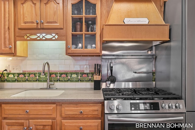 kitchen with stainless steel gas range oven, glass insert cabinets, light countertops, decorative backsplash, and a sink