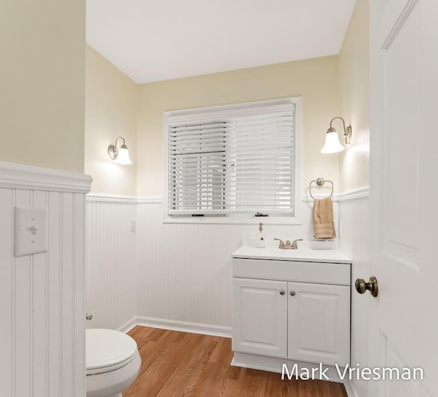 half bath with vanity, toilet, wood finished floors, and a wainscoted wall