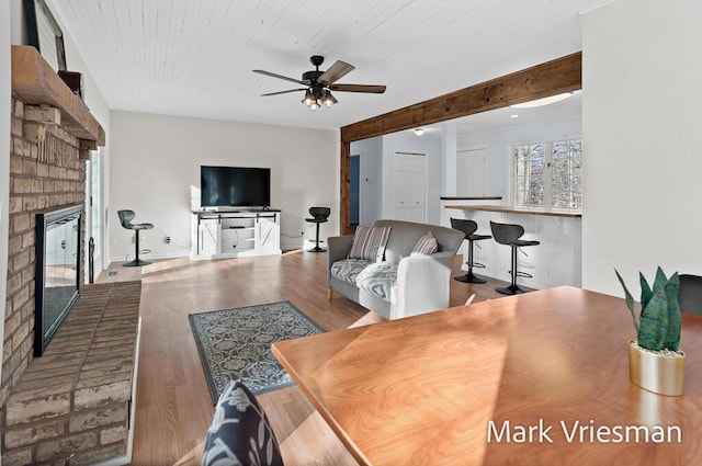 living area featuring a ceiling fan, a brick fireplace, wood finished floors, and beamed ceiling