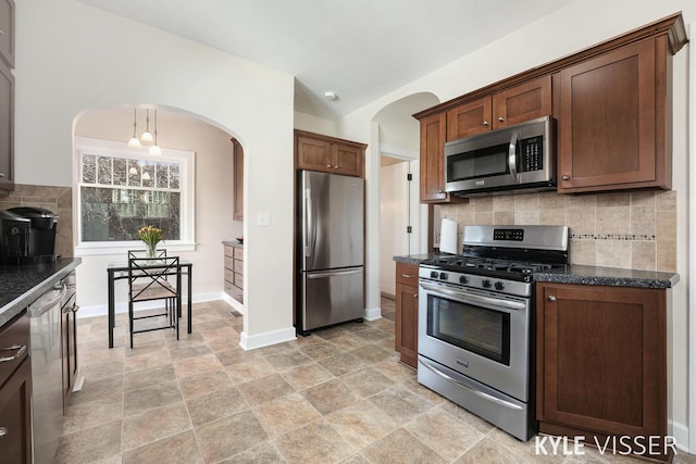 kitchen with dark stone countertops, arched walkways, appliances with stainless steel finishes, decorative backsplash, and baseboards