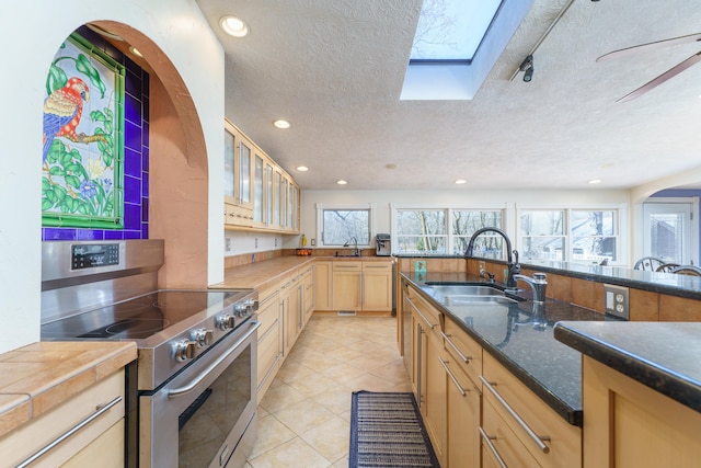 kitchen with light tile patterned floors, a sink, light brown cabinetry, stainless steel range with electric cooktop, and glass insert cabinets