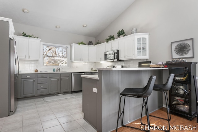 kitchen featuring a peninsula, gray cabinetry, glass insert cabinets, appliances with stainless steel finishes, and a kitchen bar