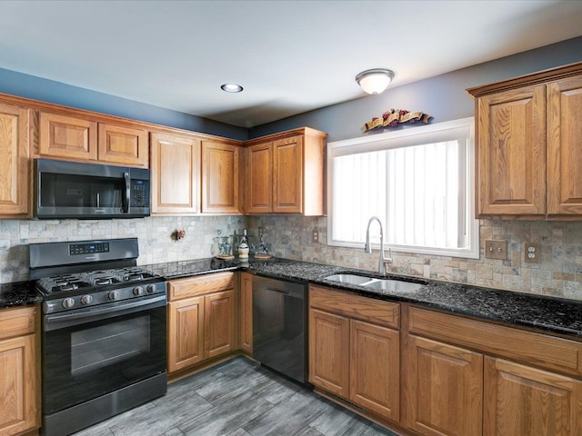 kitchen featuring brown cabinetry, dark stone countertops, black appliances, and a sink