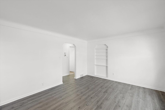 spare room featuring arched walkways, built in features, dark wood finished floors, and baseboards