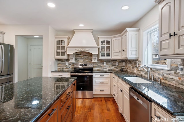 kitchen with premium range hood, a sink, wood finished floors, stainless steel appliances, and glass insert cabinets