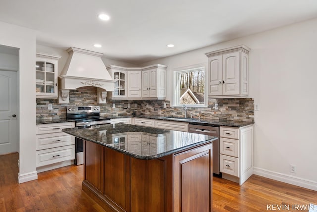 kitchen featuring wood finished floors, custom exhaust hood, a sink, stainless steel appliances, and glass insert cabinets